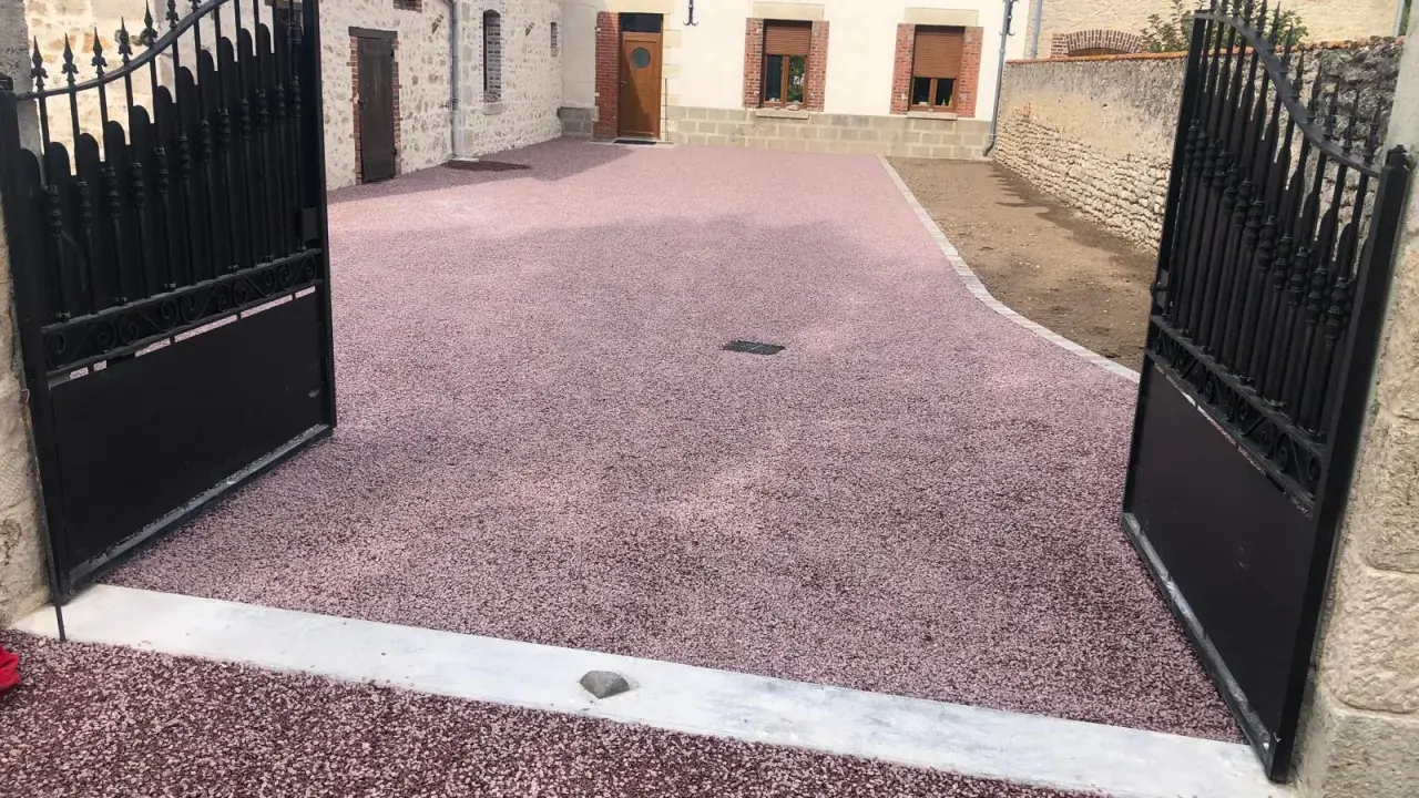 Une allée en pente pour voiture, recouverte de gravier rose, mène à une maison de campagne avec des portails en fer forgé ouverts.
