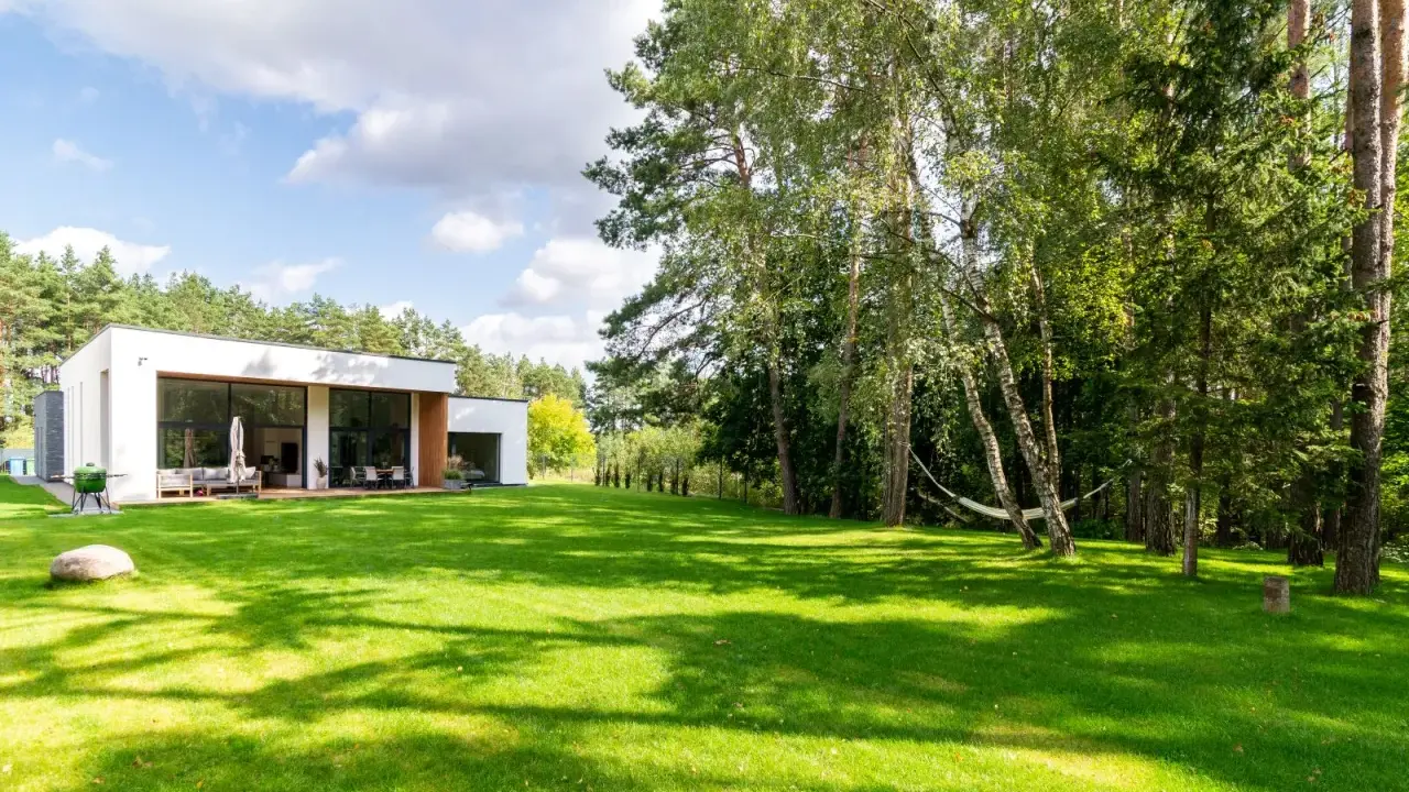 Maison moderne avec pelouse verte luxuriante et arbres, sous un ciel bleu nuageux.