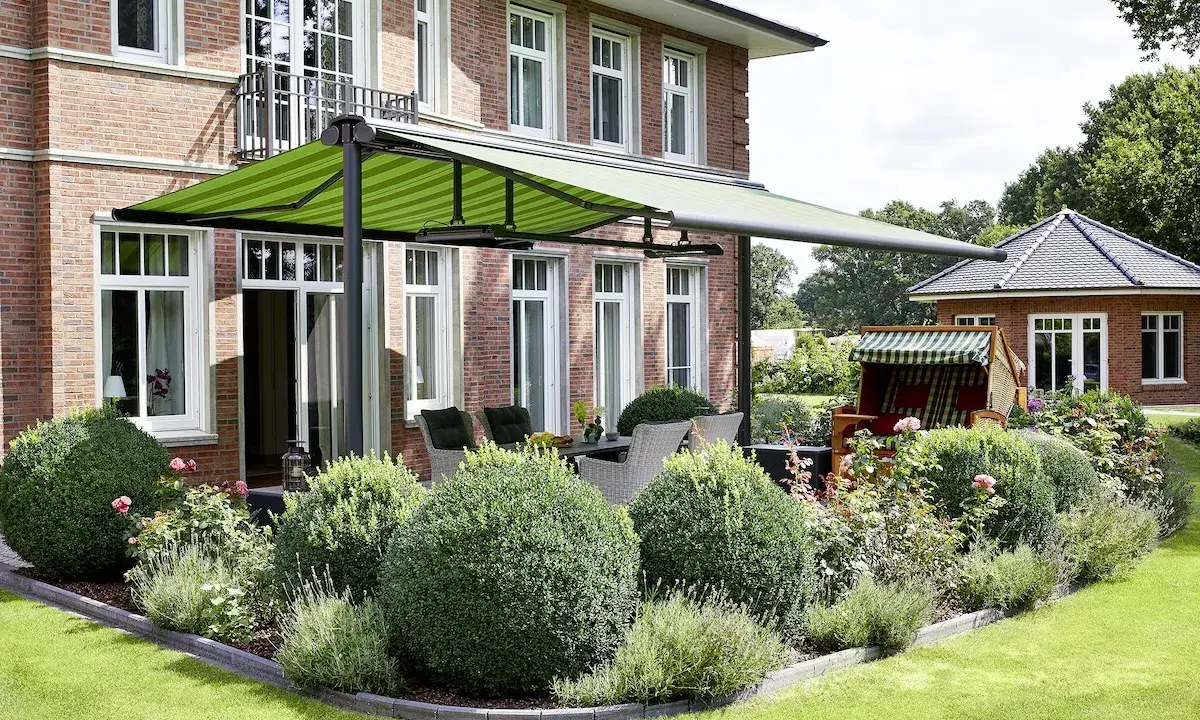 Aménagement terrasse devant maison avec auvent vert rayé, mobilier de jardin et végétation luxuriante.