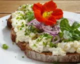 Tartine de pain noir garnie de fromage frais, oignons rouges, ciboulette et une fleur comestible orange vif.