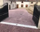 Une allée en pente pour voiture, recouverte de gravier rose, mène à une maison de campagne avec des portails en fer forgé ouverts.