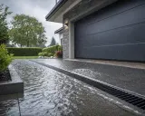 Pluie sur une allée de garage drainante, l'eau s'écoule dans la grille. La porte de garage est fermée.