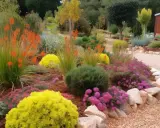 Un jardin coloré avec des fleurs orange, jaunes et roses, bordé de pierres et de paillage minéral.