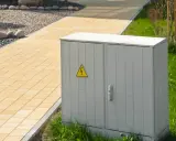 Armoire électrique grise avec symbole de danger électrique, classée IP 66, située près d'un chemin pavé jaune et d'une pelouse verte.
