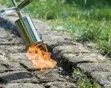 Un chalumeau brûle les mauvaises herbes entre les pavés pour empêcher leur pousse. La flamme orange vif illumine la scène.