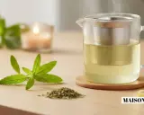 Une théière en verre remplie d'une infusion dorée, avec une branche de verveine fleur et une tranche de citron. Une bougie allumée en arrière-plan.
