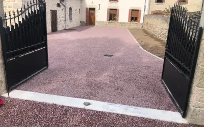 Une allée en pente pour voiture, recouverte de gravier rose, mène à une maison de campagne avec des portails en fer forgé ouverts.