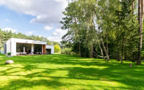Maison moderne avec pelouse verte luxuriante et arbres, sous un ciel bleu nuageux.