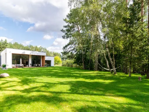 Maison moderne avec pelouse verte luxuriante et arbres, sous un ciel bleu nuageux.