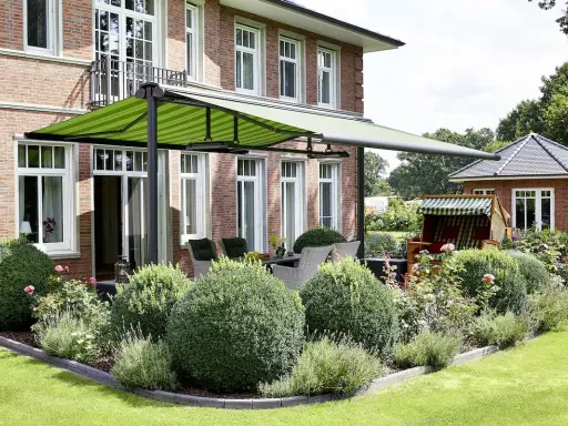 Aménagement terrasse devant maison avec auvent vert rayé, mobilier de jardin et végétation luxuriante.