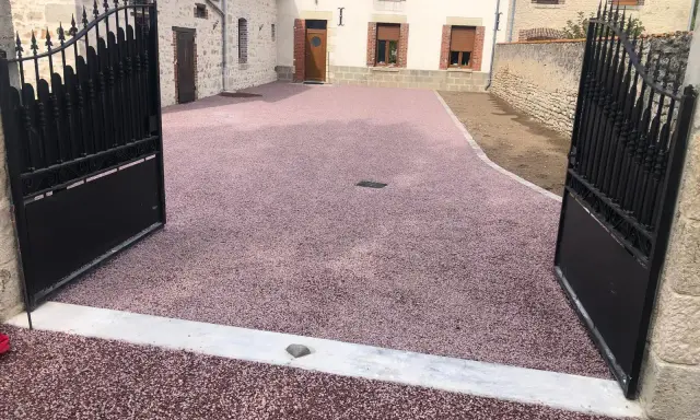 Une allée en pente pour voiture, recouverte de gravier rose, mène à une maison de campagne avec des portails en fer forgé ouverts.