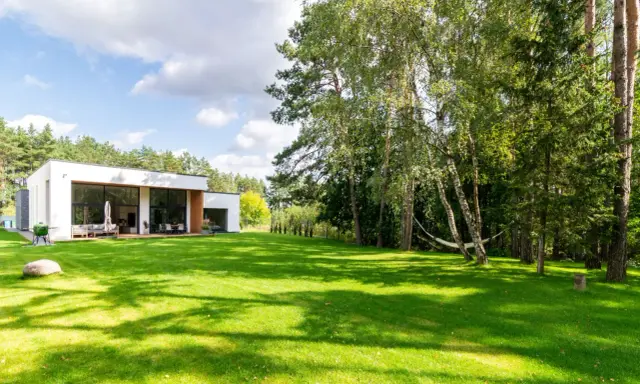 Maison moderne avec pelouse verte luxuriante et arbres, sous un ciel bleu nuageux.