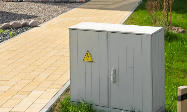 Armoire électrique grise avec symbole de danger électrique, classée IP 66, située près d'un chemin pavé jaune et d'une pelouse verte.