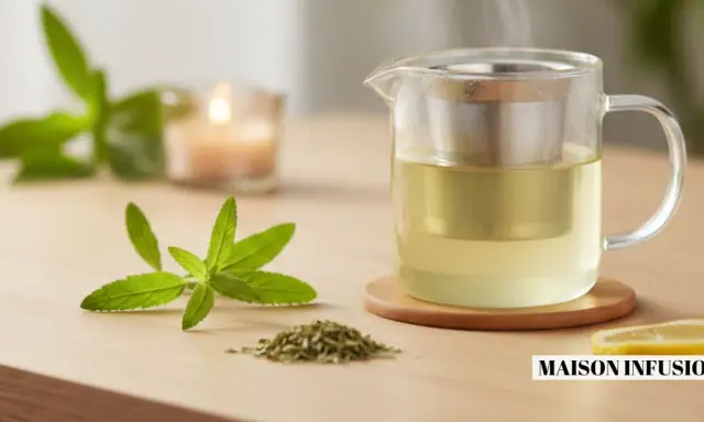 Une théière en verre remplie d'une infusion dorée, avec une branche de verveine fleur et une tranche de citron. Une bougie allumée en arrière-plan.