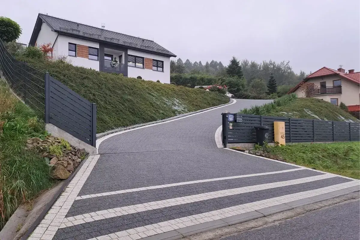 Une allée en pente pour voiture serpente vers une maison moderne, bordée de végétation luxuriante et d'une clôture sombre.