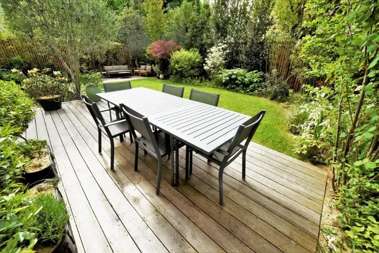 Table et chaises sur une terrasse en bois, entourées d'un jardin verdoyant. Un bel aménagement extérieur pour profiter des beaux jours.