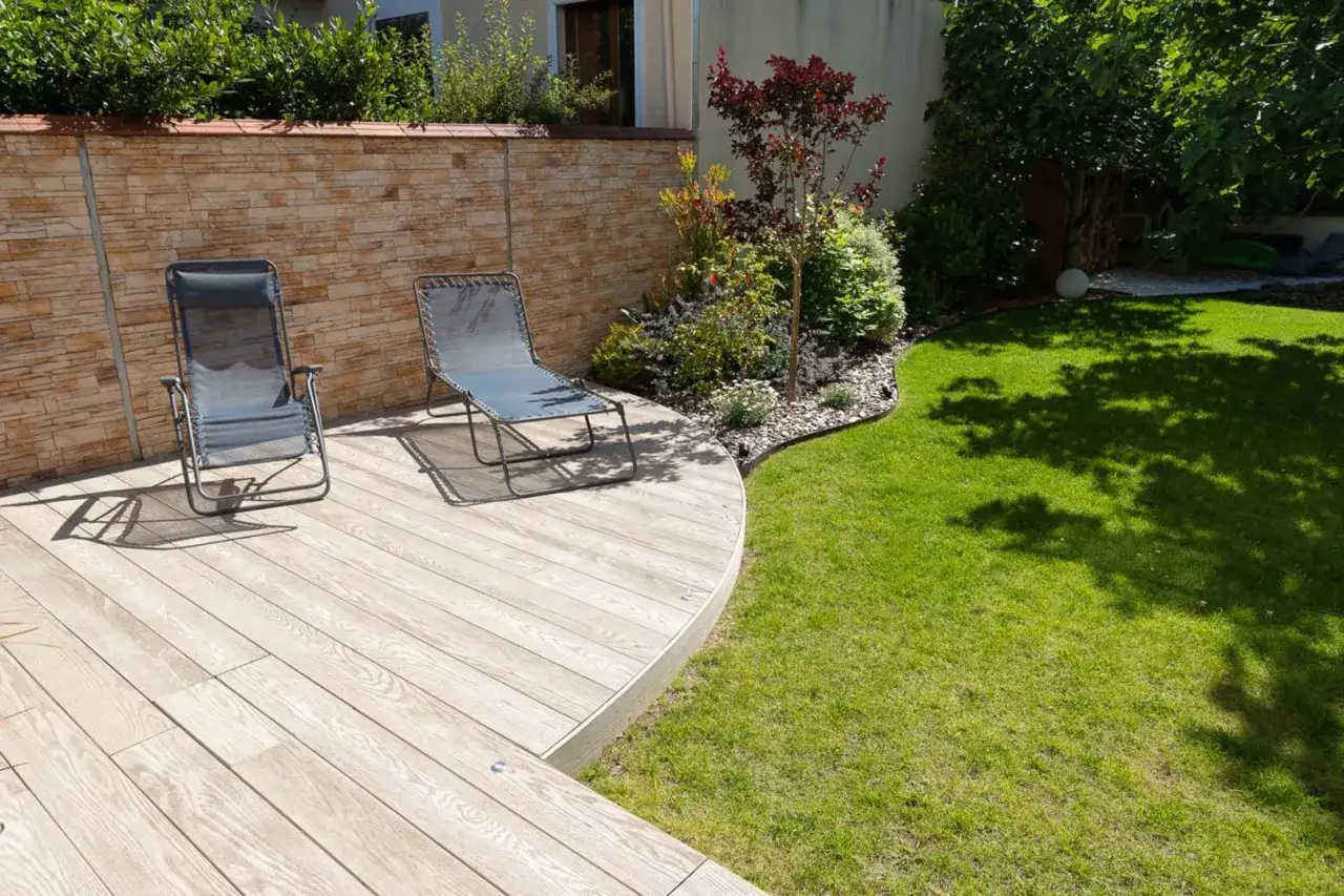 Aménagement extérieur : terrasse en bois, deux chaises longues, mur de pierres et pelouse verdoyante. Un espace détente parfait.