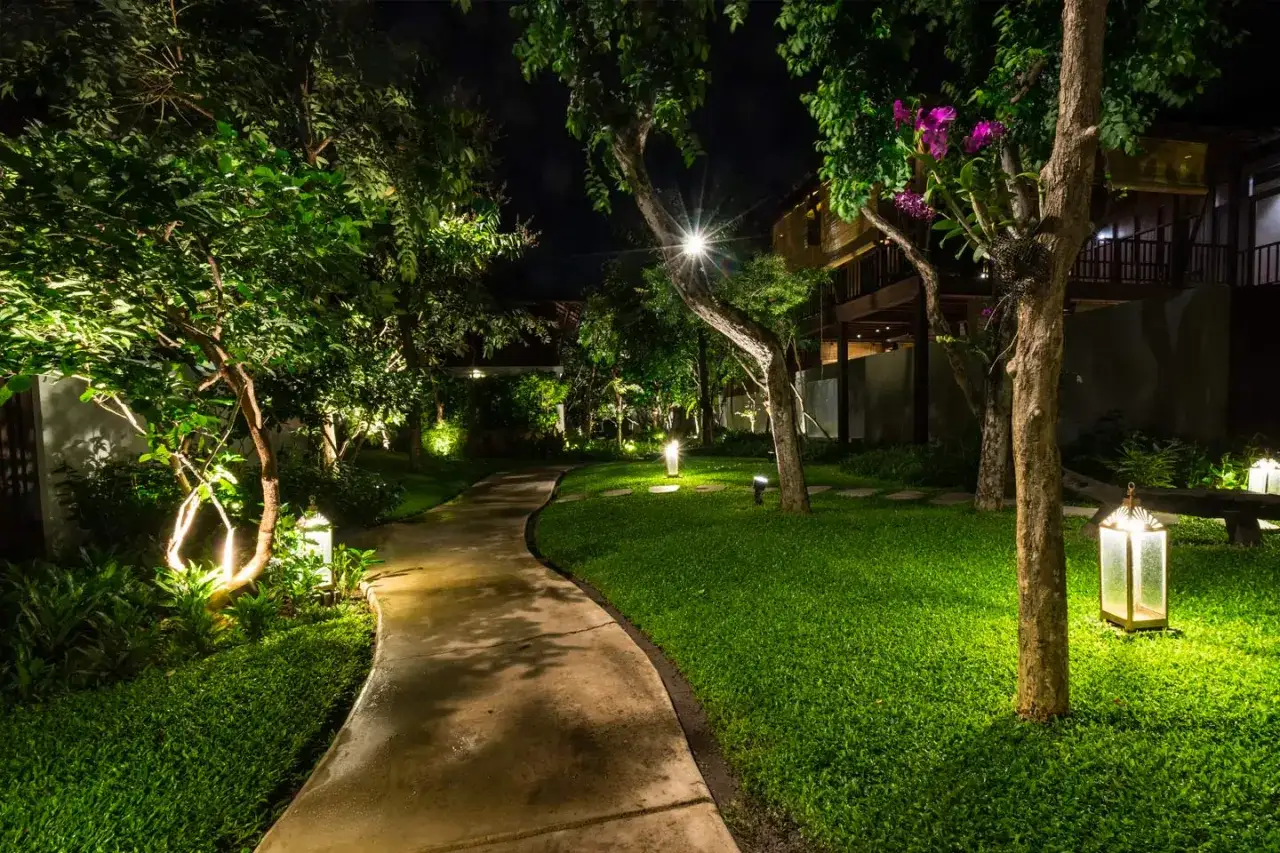 Sentier sinueux éclairé la nuit, bordé d'une pelouse verte et d'arbres. La lumière douce, de couleur blanc neutre, crée une atmosphère paisible.