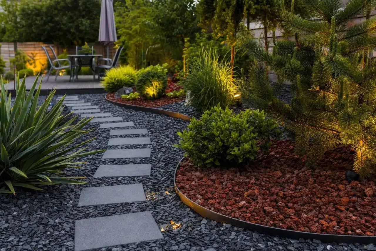 Allée de dalles grises sur un lit de gravier noir, bordée de paillage minéral rouge. Plantes vertes et éclairage d'ambiance.