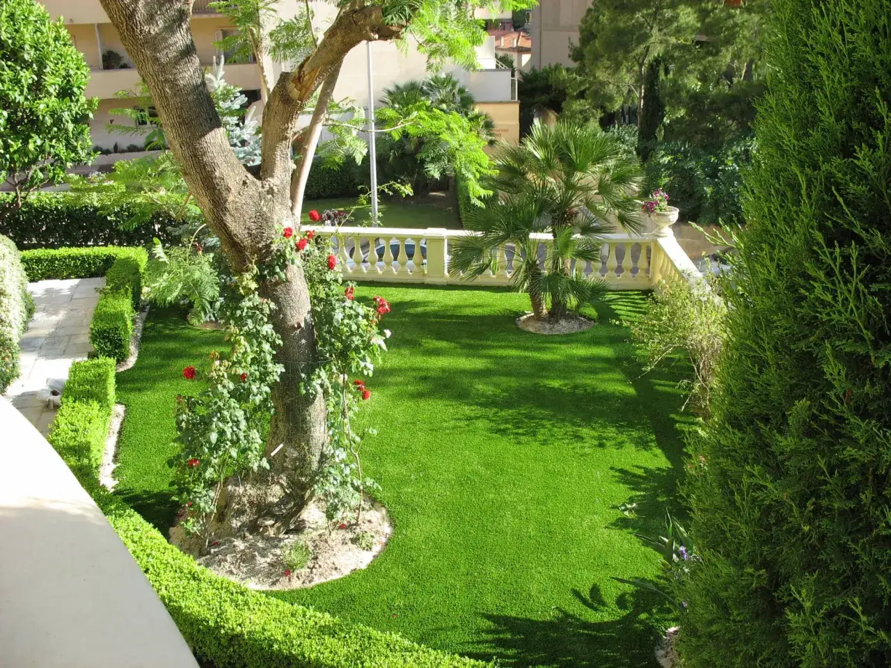 Un jardin luxuriant avec du gazon synthétique parfait, idéal pour savoir comment choisir son gazon synthétique. Un arbre imposant et des rosiers ajoutent du charme.
