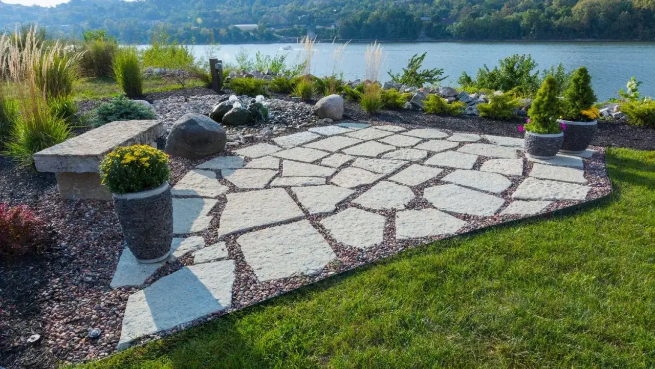 Une belle terrasse en pavé irrégulier, bordée de gravier, avec un banc en pierre et des plantes, surplombant une rivière paisible.