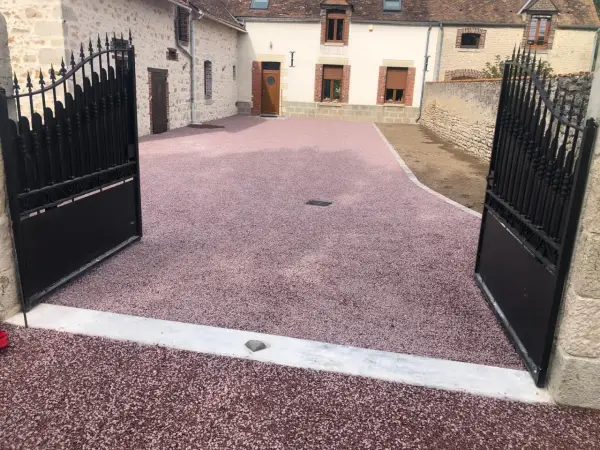Une allée en pente pour voiture, recouverte de gravier rose, mène à une maison de campagne avec des portails en fer forgé ouverts.